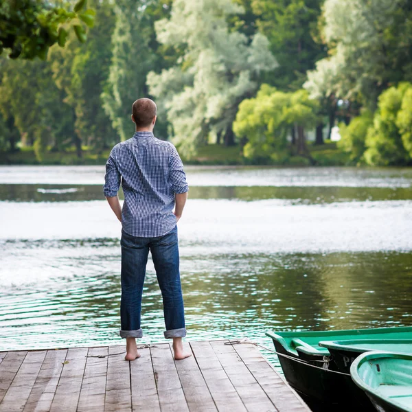 Young handsome man standing on wooden pier, relaxing and meditaing in summer morning, back view