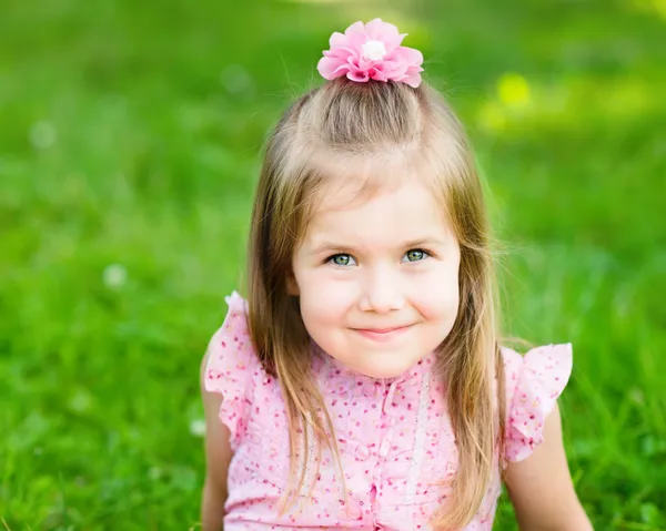 Sweet smiling little girl with long blond hair, sitting on grass in summer park, closeup outdoor portrait