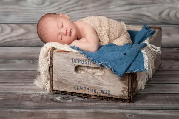 One week old newborn baby boy sleeping on his stomach in a vintage, wooden soda pop crate lined with frayed burlap and denim.