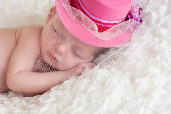 A portrait of a sleeping newborn baby girl wearing a fancy, pink, brimmed hat with netting over her face.