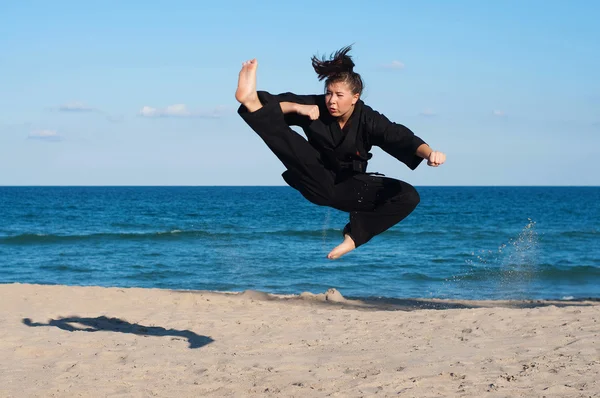 Female, fourth degree, Taekwondo black belt athlete performs a midair jumping kick on the beach.