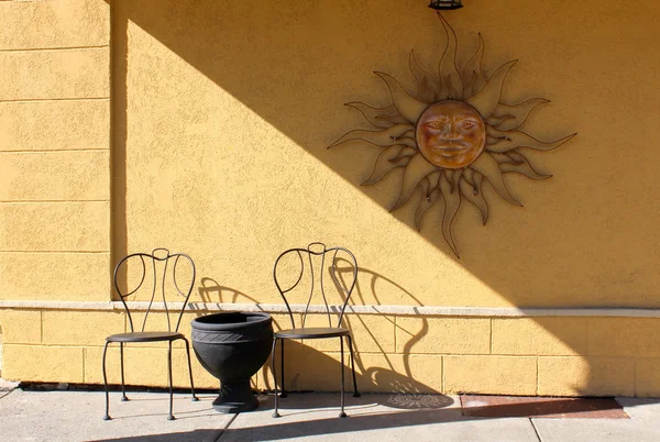 Yellow painted stucco wall with old rustic metal sun decoration hanging over black metal chairs and table at outdoor eatery