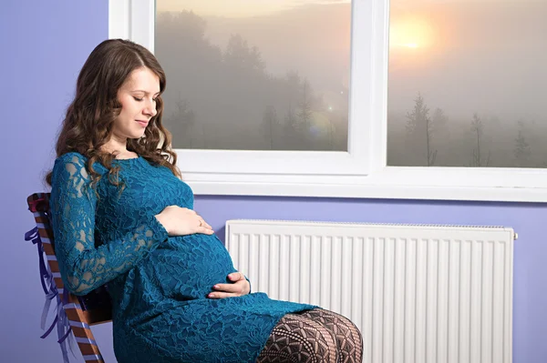 The pregnant girl is sitting on the chair near the window