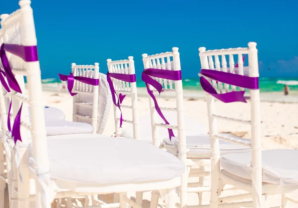 White wedding chairs decorated with purple bows on sandy beach