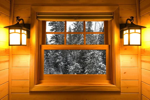 Inside view snow covered trees in a cedar cabin window with lights on