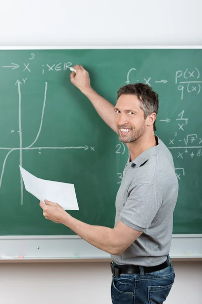 Teacher Holding Paper While Writing On Chalkboard