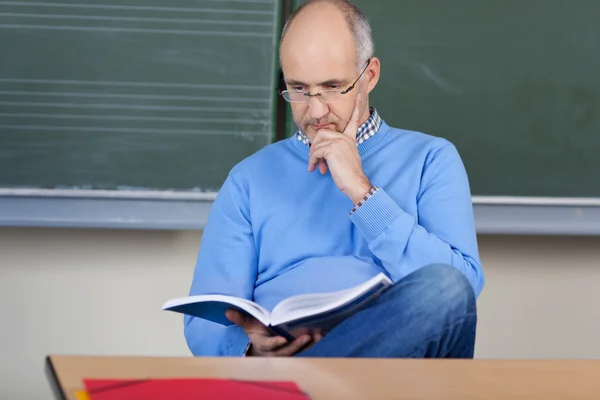 Professor Reading Book At Desk In Classroom