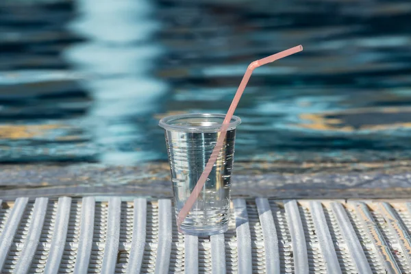 A plastic cup with drink and pink straw standing near the swimming pool edge