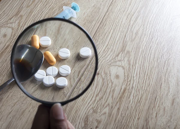 Syringe, pills and depiction of heroin seen through magnifying glass on wooden board