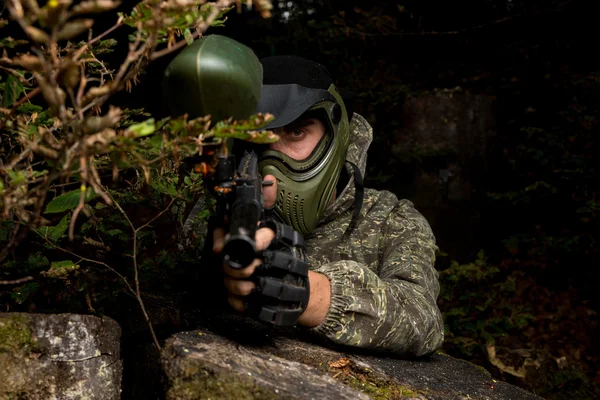 Paintball sport player in protective uniform and mask aiming and shooting with gun outdoors