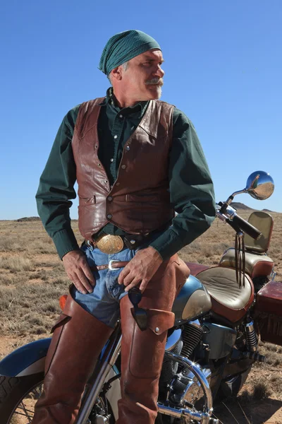 Handsome older retired man in desert leaning against motorcycle staring off into the distance