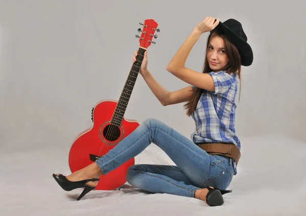 Young girl sitting in a blouse with short sleeves, his right hand on the neck of the red guitar , and his left hand on the head of a black hat