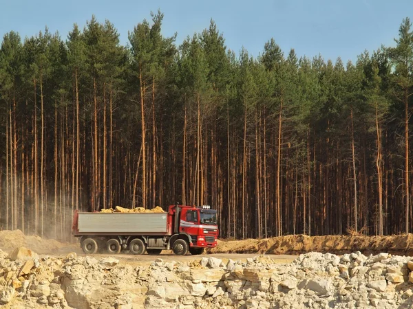 Big truck with red cabin and gray semitrailer full of loaded material. Open mine in pine forest