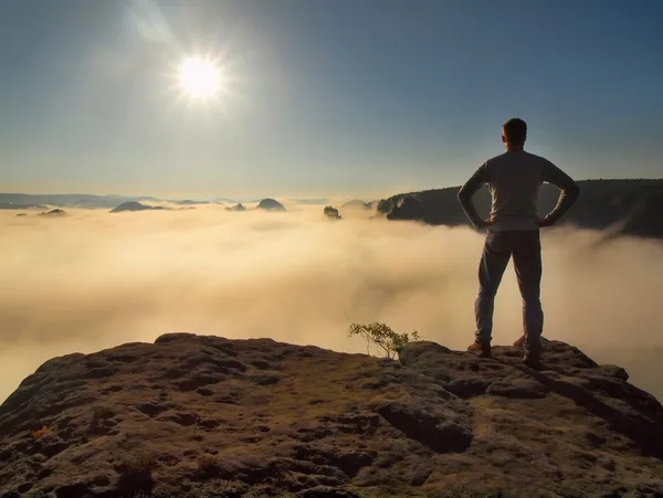 Man in shirt and jeans is standing on the peak of sandstone rock in national park Saxony Switzerland and watching over the misty and foggy morning valley to Sun. Beautiful moment the miracle of nature