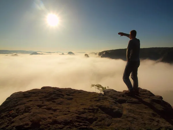 Tourist in jeans is standing on the peak of sandstone rock in national park Saxony Switzerland and watching over the morning misty valley to big Sun.