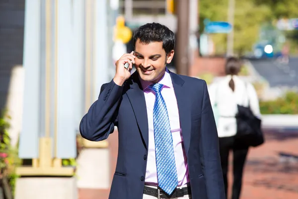 A closeup portrait of a young busy, happy, smiling business man talking on his phone while walking outside down the street holding a briefcase