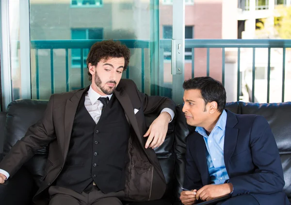 Closeup portrait of two guys in business suits sitting on black couch. One is crying about life problems and other probably caused the trouble or is indifferent