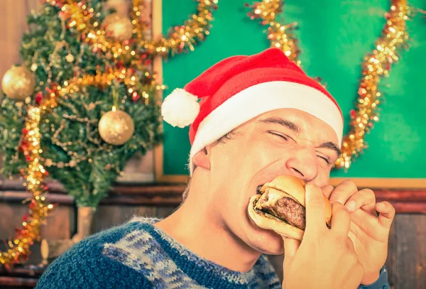 Portrait of an handsome young man with Santa hat eating cheeseburger