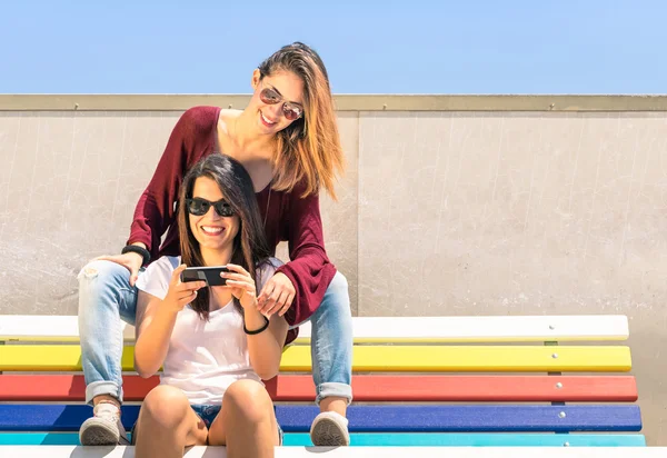 Best friends enjoying time together outdoors with smartphone - Concept of new technology with two girlfriends having fun on a multicolored bench