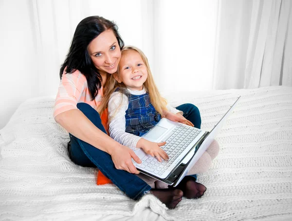 Happy mother and daughter working on computer sitting on bed together