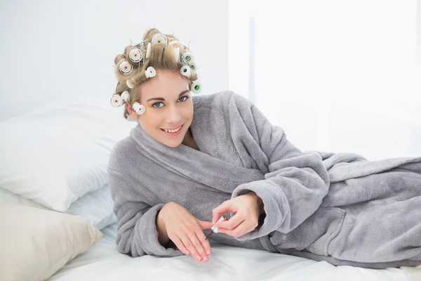 Delighted relaxed blonde woman in hair curlers applying nail polish