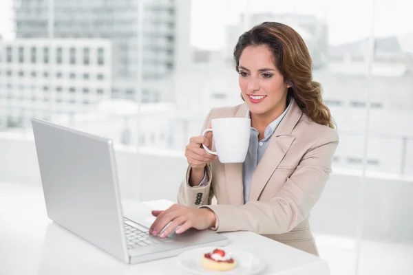 Cheerful businesswoman using laptop at her desk and holding mug