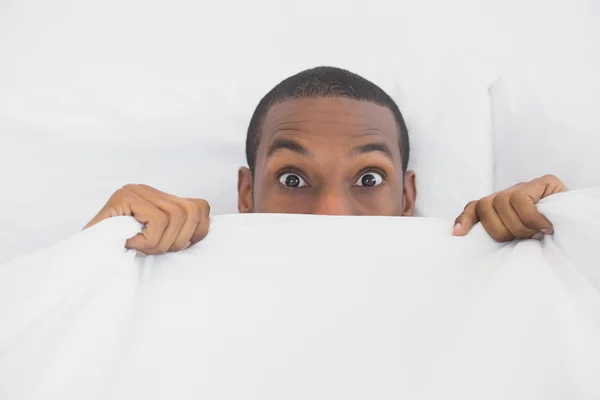 Shocked Afro man covering face with sheet in bed