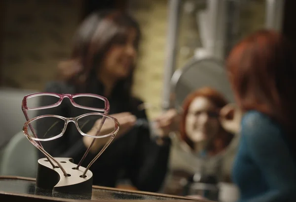 Glasses Frames On Display With Woman Trying On Glasses With Help In The Background