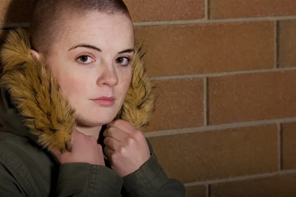 Teen Girl With Shaved Head Holding Onto Hood Of Coat And Leaning On Brick Wall