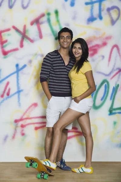 Couple standing with a skateboard in front of a graffiti covered wall