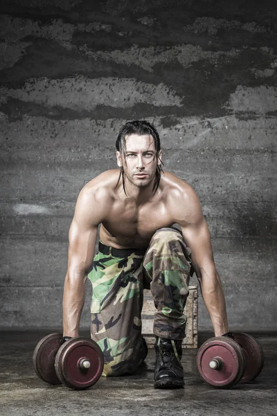 Muscle athlete with camouflage pants looking at camera and weigh tlifting