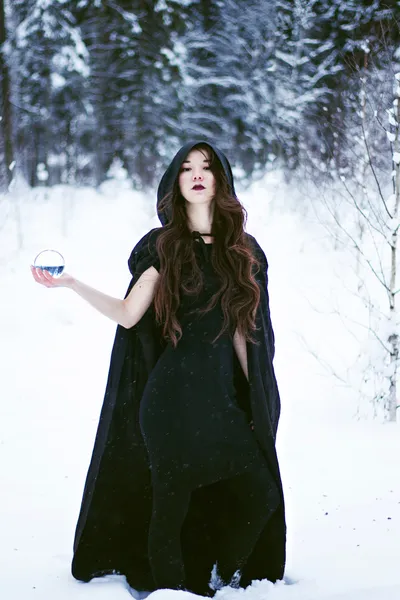 Witch or woman in black cloak with glass ball in white snow forest