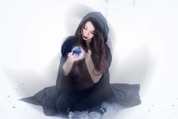 Witch or woman doing magic in black cloak with glass ball in white snow forest