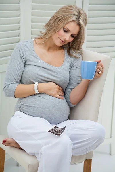 Attractive pregnant woman sitting at home on chair holding blue mug, smiling and touching her tummy