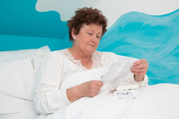 Portrait: ill senior woman reading paper of side effects in bed.