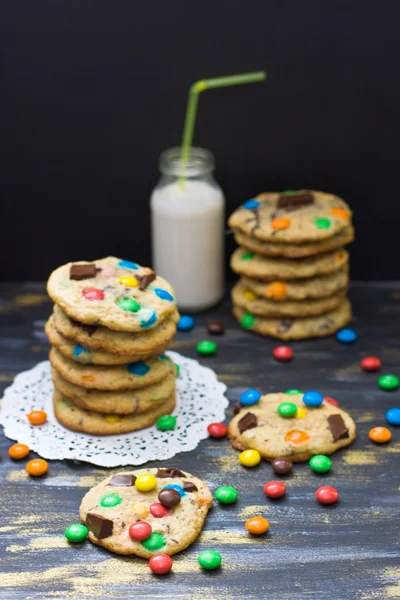 Cookies with colorful candy pieces, coarsly chopped chocolate with bottle of milk on dark backgroond