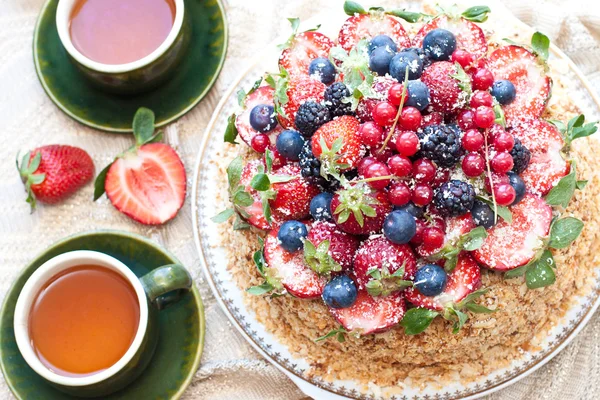 Napoleon cake, decorated with fresh berries, strawberries, blueberries and blackberries, with birthday candle. Two green tea cups and teapot near the window.