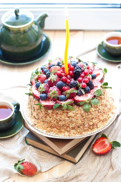 Napoleon cake, decorated with fresh berries, strawberries, blueberries and blackberries, with birthday candle. Two green tea cups and teapot near the window.