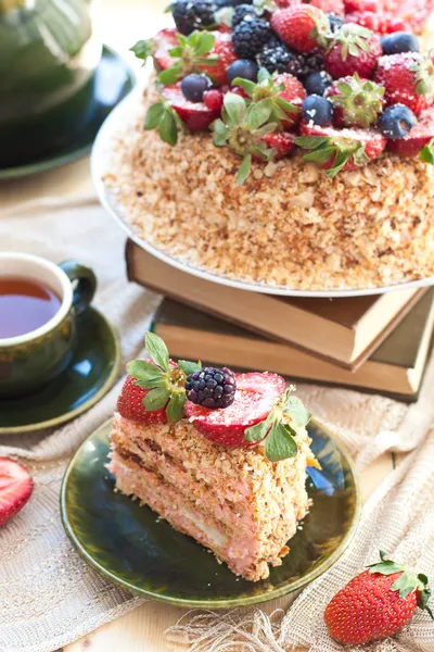 Napoleon cake, decorated with fresh berries, strawberries, blueberries and blackberries, with birthday candle. Two green tea cups and teapot near the window.