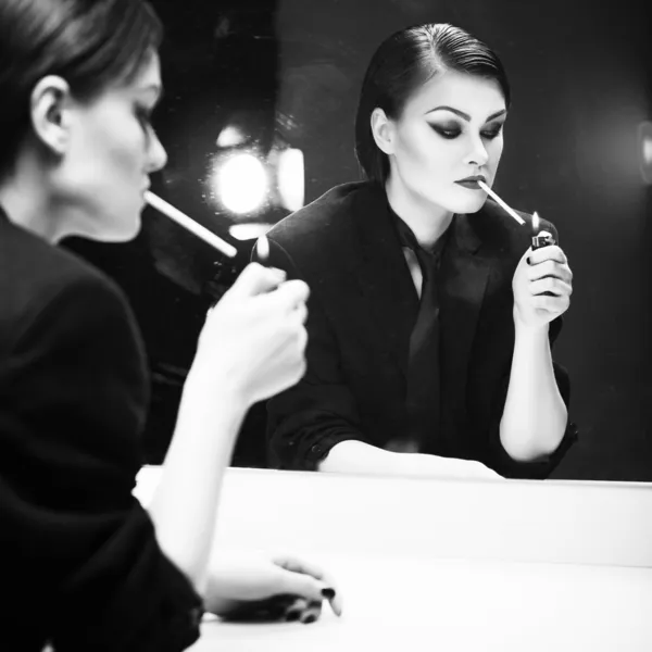 Beautiful brunette woman in business costume lighting a cigarette in front of the mirror with color backlight. Black and White