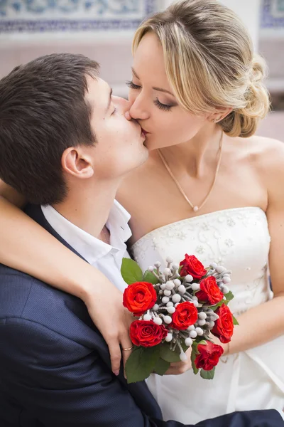 Young couple kissing in wedding gown. Bride holding bouquet with red roses