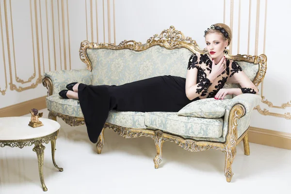Beautiful blonde royal woman laying on a retro sofa in gorgeous luxury dress. Indoor. Copy Space