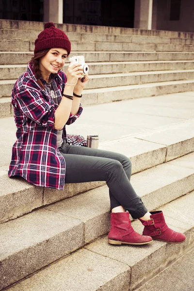 Brunette woman in hipster outfit sitting on steps on the street, photographing. Toned image