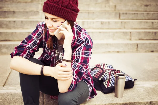 Brunette woman in hipster outfit sitting on steps and talking on the phone on the street. Toned image. Copy Space