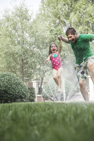Father holding daughters hand while she jumps through the sprinkler