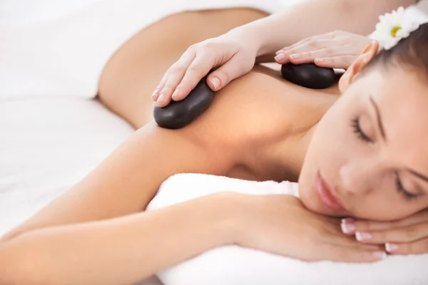 Hot stone massage. Beautiful young woman lying on front while massage therapist massaging her back with spa stones