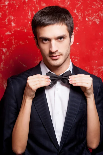 Man in formalwear standing against red background while woman adjusting his bow tie
