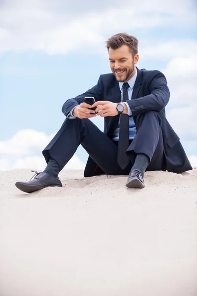 Businessman looking at mobile phone sitting on sand
