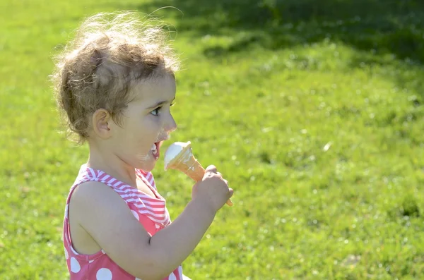 Pretty young girl eating ice cream. She is wearing a pink and white dress, with the sun behind her (backlit). She has blonde curly hair and is looking towards the camera.