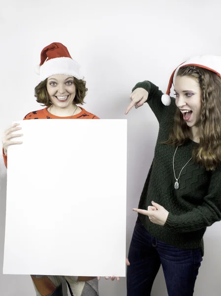 Two pretty young women wearing Santa hats and holding blank sign for copy space.